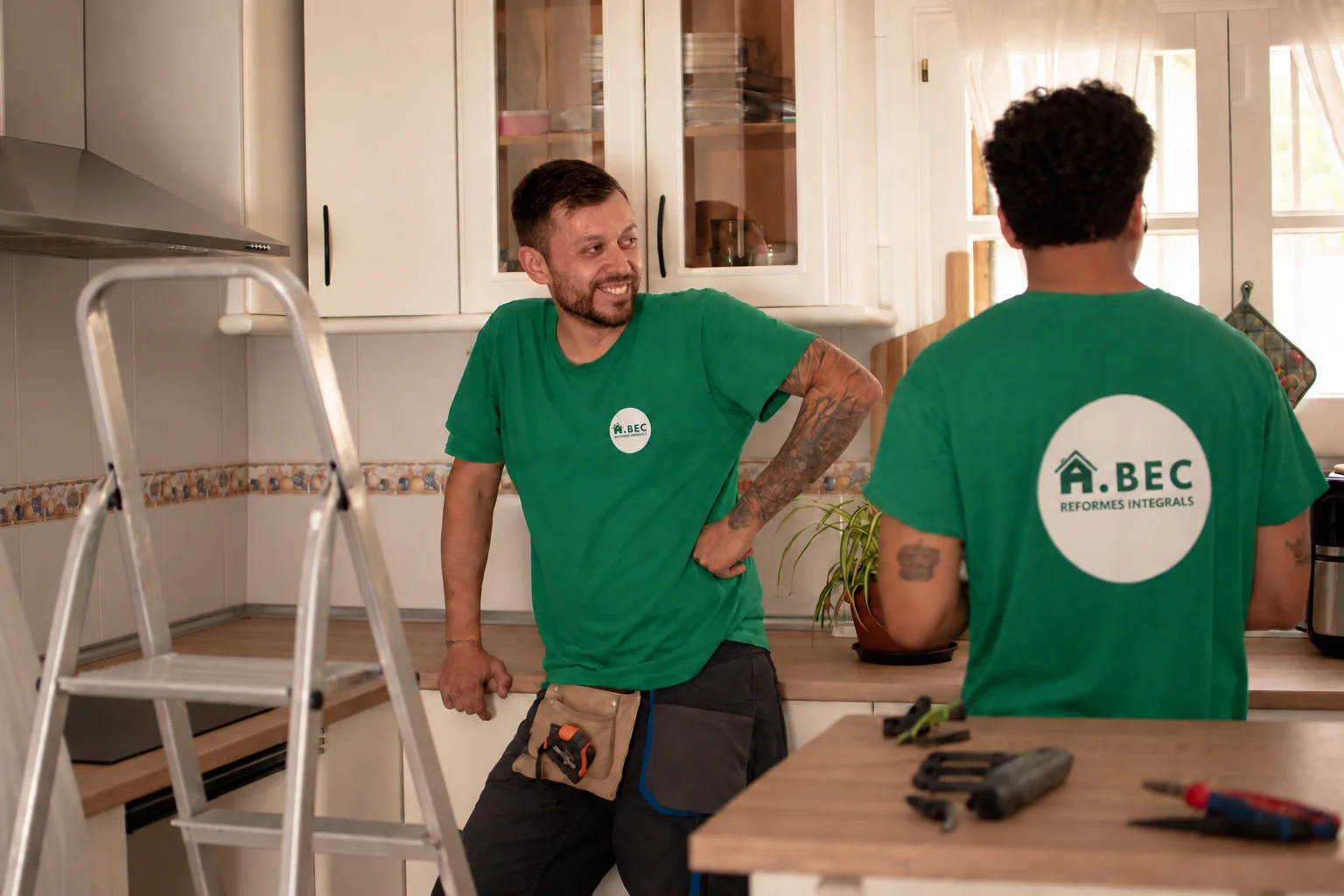 Dos membres de l’equip d’A.BEC conversant en una cuina, al costat d’una escala i eines de treball.